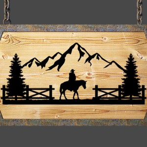 May include: A wooden sign with a rustic brown finish, featuring a silhouette of a cowboy riding a horse in front of a mountain range and two pine trees. The sign is hanging from two chains.