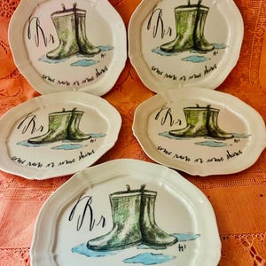 Może przedstawiać: Zestaw pięciu białych ceramicznych talerzy z falistym brzegiem. Każdy talerz przedstawia ilustrację zielonych kaloszy z napisem "Rr" i frazą "come rain or come shine". Talerze mają fantazyjny, artystyczny design.