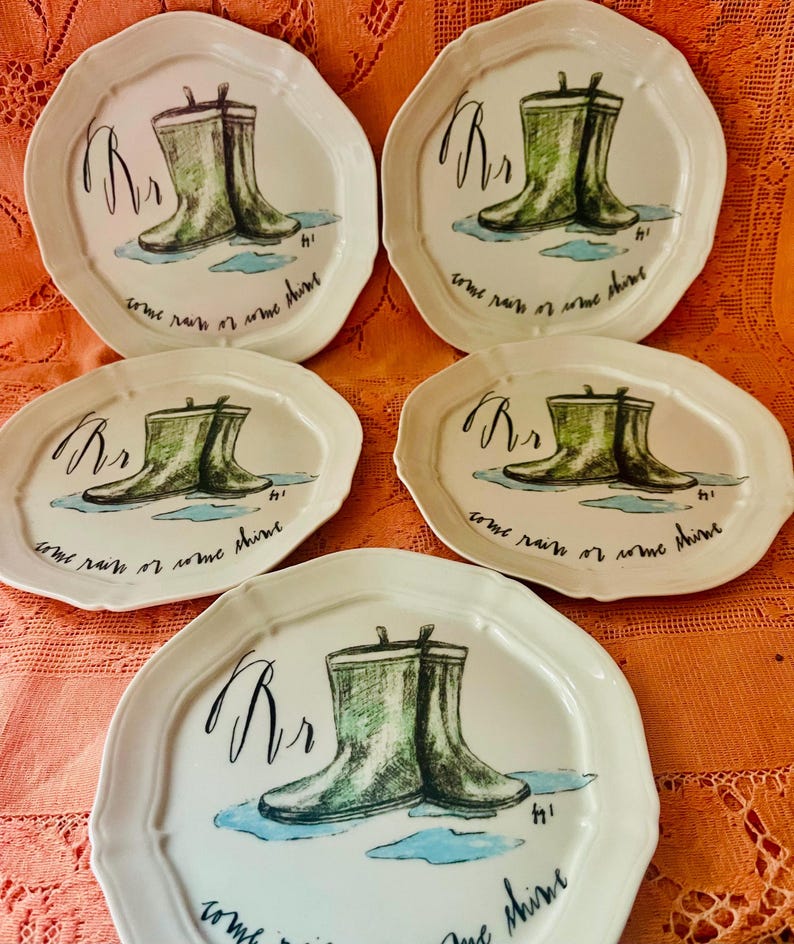 Może przedstawiać: Zestaw pięciu białych ceramicznych talerzy z falistym brzegiem. Każdy talerz przedstawia ilustrację zielonych kaloszy z napisem "Rr" i frazą "come rain or come shine". Talerze mają fantazyjny, artystyczny design.