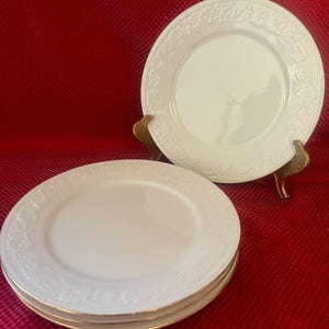 May include: Four white ceramic dinner plates with a gold rim and an embossed floral design.