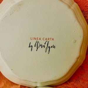 Może przedstawiać: Zbliżenie białego ceramicznego talerza z falistą krawędzią. Talerz zawiera tekst "LINEA CARTA" w kolorze czerwonym i "by Diva Iyari" w czarnym skrypcie. Talerz znajduje się na teksturowanej powierzchni.