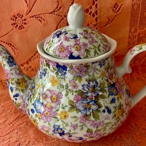 May include: A white ceramic teapot with a floral pattern in shades of pink, blue, yellow, and purple. The teapot has a curved spout, a rounded body, and a handle. The lid is topped with a white, tulip-shaped finial.