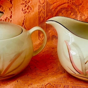 May include: Cream-colored sugar bowl with lid and creamer set. The sugar bowl has a dark brown finial and handles. Both pieces are decorated with pink and brown leaf-like designs.