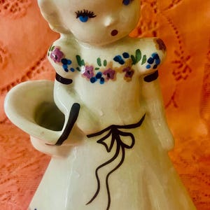 May include: A vintage ceramic figurine of a young girl holding a basket. The figurine is painted in cream and adorned with floral details in blue, purple, and green. The girl has blue eyes and a delicate expression.