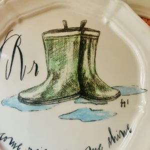 Może przedstawiać: Biały ceramiczny talerz z falistym brzegiem, z zieloną ilustracją kaloszy w kałuży. Talerz ma napis "come rain or come shine" i literę "Rr".