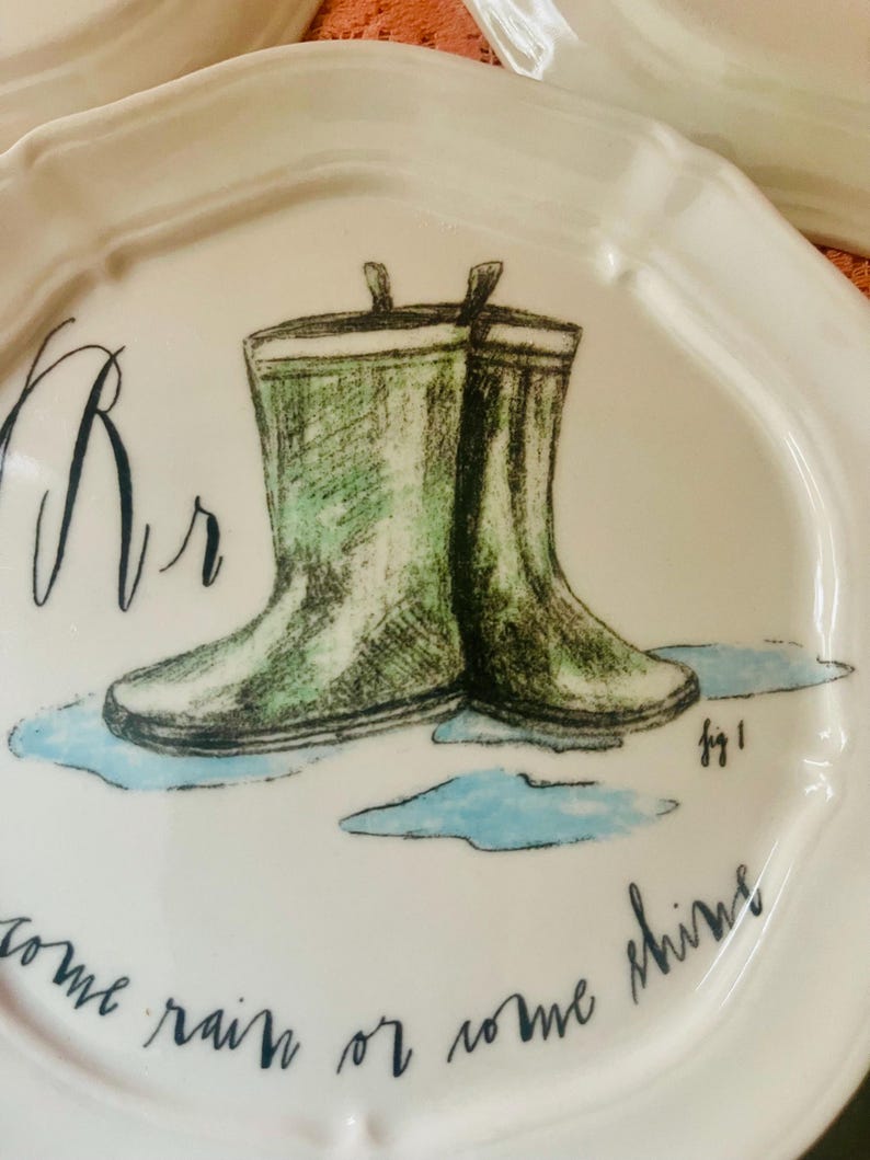 Może przedstawiać: Biały ceramiczny talerz z falistym brzegiem, z zieloną ilustracją kaloszy w kałuży. Talerz ma napis "come rain or come shine" i literę "Rr".