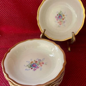 May include: A stack of off-white porcelain bowls with a scalloped gold rim and a floral center design. The bowls are stacked on a red surface. One bowl is displayed on a decorative stand.