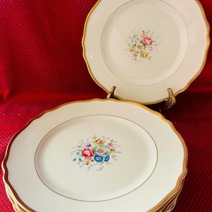 May include: A set of six white ceramic dinner plates with a gold rim and a floral design. The plates have a scalloped edge and are stacked on top of each other.