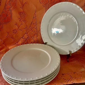 May include: A stack of white ceramic plates with a beaded edge, and two additional plates. The plates are on an orange tablecloth. The plates are a simple, elegant design.