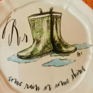 Może przedstawiać: Biały ceramiczny talerz z zieloną akwarelową ilustracją kaloszy. Talerz ma literę "Rr" i frazę "come rain or come shine" napisaną czarnym pismem. Talerz ma falisty brzeg.