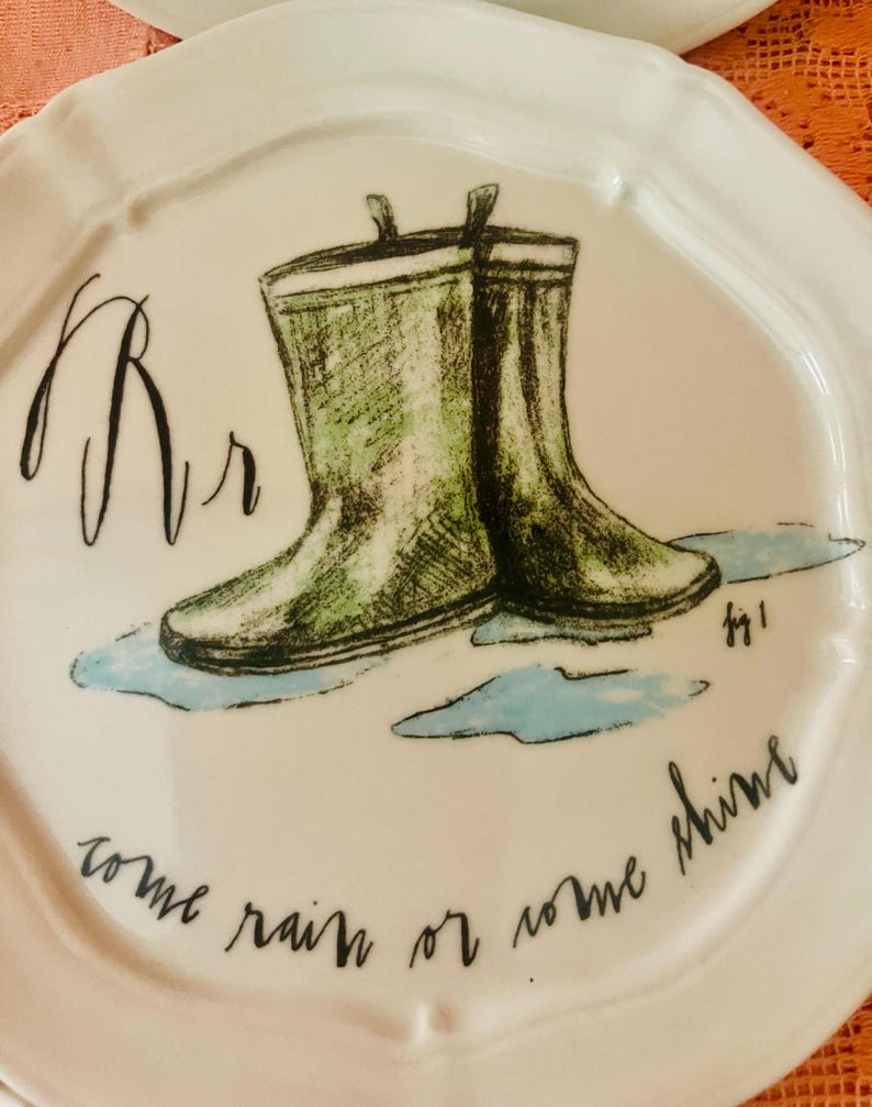 Może przedstawiać: Biały ceramiczny talerz z zieloną akwarelową ilustracją kaloszy. Talerz ma literę "Rr" i frazę "come rain or come shine" napisaną czarnym pismem. Talerz ma falisty brzeg.