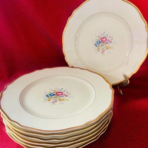 May include: A stack of eight white dinner plates with a gold rim and a floral design in the center. The plates have a scalloped edge.