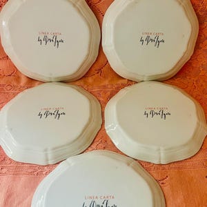 Może przedstawiać: Zestaw pięciu białych ceramicznych talerzy z falistym brzegiem. Na spodzie każdego talerza znajduje się czarny napis "LINEA CARTA by Pr&eacute;szen". Talerze są ułożone na teksturowanej, brzoskwiniowej powierzchni.