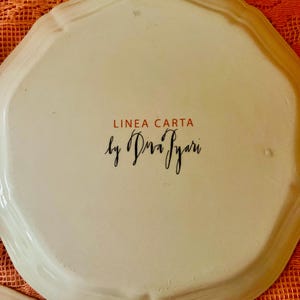 Może przedstawiać: Kremowy ceramiczny talerz z falistą krawędzią. Tekst "LINEA CARTA" jest czerwony, a "by Diva Iyari" jest czarną czcionką. Talerz leży na pomarańczowej, teksturowanej powierzchni.