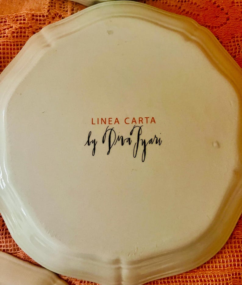 Może przedstawiać: Kremowy ceramiczny talerz z falistą krawędzią. Tekst "LINEA CARTA" jest czerwony, a "by Diva Iyari" jest czarną czcionką. Talerz leży na pomarańczowej, teksturowanej powierzchni.