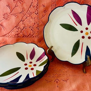 May include: White ceramic plates with a scalloped edge and a floral design. The plates feature a white background with a colorful flower design in purple, green, red, and yellow. The plates have a dark blue rim and are on a pink tablecloth.