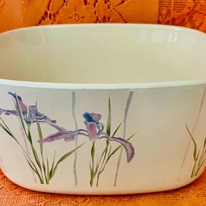 May include: Off-white oval casserole dish with two handles. The dish features a floral design of purple irises and green stems. The dish is likely made of ceramic or glass.