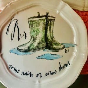 Może przedstawiać: Biały ceramiczny talerz z falistym brzegiem, z ręcznie rysowaną ilustracją zielonych kaloszy w kałuży. Talerz ma literę "Rr" i frazę "come rain or come shine" w czarnym skrypcie.