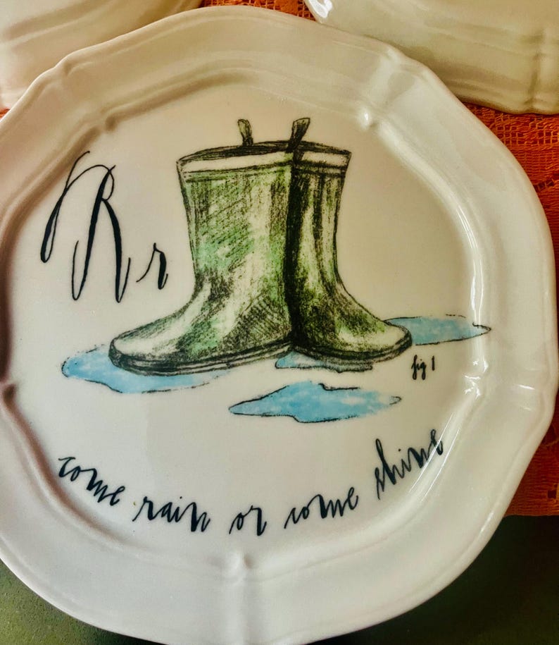 Może przedstawiać: Biały ceramiczny talerz z falistym brzegiem, z ręcznie rysowaną ilustracją zielonych kaloszy w kałuży. Talerz ma literę "Rr" i frazę "come rain or come shine" w czarnym skrypcie.
