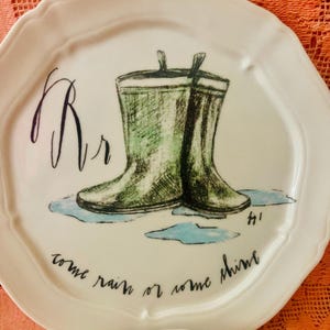 Może przedstawiać: Biały ceramiczny talerz z zieloną ilustracją kaloszy. Talerz zawiera napis "come rain or some shine" i literę "Rr". Talerz ma falisty brzeg.