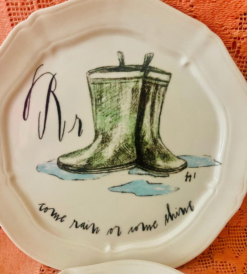 Może przedstawiać: Biały ceramiczny talerz z zieloną ilustracją kaloszy. Talerz zawiera napis "come rain or some shine" i literę "Rr". Talerz ma falisty brzeg.