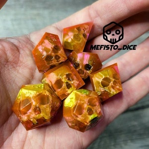 Può includere: Set di sette dadi iridescenti con un design a forma di teschio. I dadi sono una miscela di arancione e giallo con una finitura scintillante. I dadi sono in una mano, e il testo "MEFISTO_DICE" è visibile sullo sfondo.