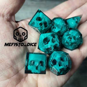 May include: Set of seven teal green dice with a skull design inside each die. The dice are on a hand.