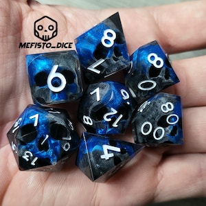 Pode incluir: Conjunto de sete dados azuis e pretos com números brancos e desenhos de caveiras. Os dados são feitos de resina e têm um aspeto salpicado.
