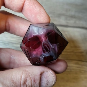 Può includere: Un dado d20 trasparente, iridescente viola e bianco con numeri neri. Il dado ha un design a forma di teschio su un lato.