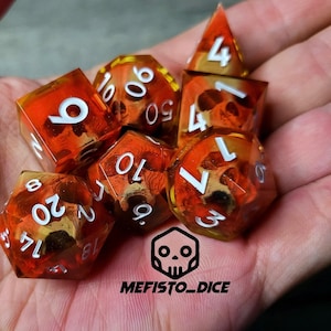 May include: Set of six orange and yellow dice with white numbers and skull designs. The dice are in a hand.