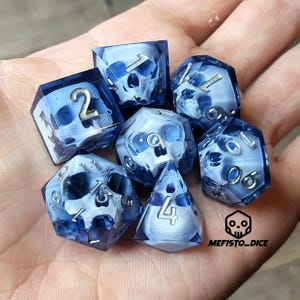Puede incluir: Conjunto de siete dados azules y blancos con un diseño de calavera. Los dados son translúcidos y tienen un acabado mate. Los números en los dados son visibles a través del material translúcido.