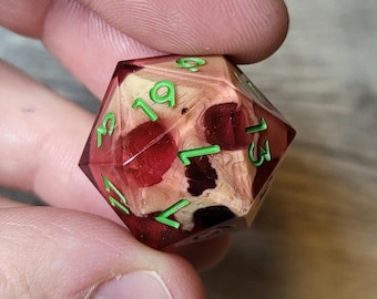 May include: A red and white skull-shaped dice with green numbers. The numbers on the dice are 1, 2, 3, 4, 5, 6, 7, 8, 9, 10, 11, 12, 13, 14, 15, 16, 17, 18, 19, 20.
