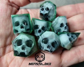 Puede incluir: Juego de siete dados poliédricos con un diseño de calavera en el interior. Los dados son transparentes con un borde verde y la calavera es negra y blanca. Los dados son para juegos de mesa y tienen el texto "MEFISTO_DICE" en la parte inferior derecha.
