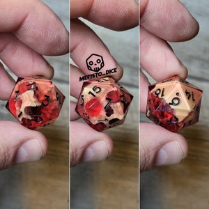 Può includere: Un dado rosso e crema a più facce con numeri neri. Il dado ha un design a forma di teschio su un lato.