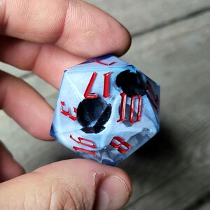 Può includere: Un dado d20 blu e bianco con numeri rossi e accenti neri. Il dado è traslucido e mostra i numeri 1, 3, 8, 10, 11 e 17.