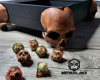 May include: A wooden box with skull-shaped legs, used for dice games. The box is open and there are several dice inside, some with green and red accents. The dice are on a wooden surface. The text "MEFISTO_DICE" is visible on the surface.