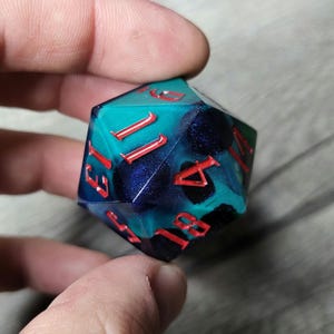 Può includere: Un dado iridescente blu e verde con numeri rossi. Il dado è un dado a 12 facce con i numeri da 1 a 12.