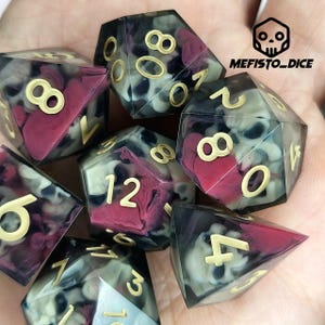 Può includere: Set di sette dadi poliedrici a tema teschio. I dadi sono trasparenti con un effetto marmorizzato nero, bianco e rosso. I numeri dorati sono visibili su ogni dado. Il testo "MEFISTO_DICE" si trova nell'angolo in alto a destra.