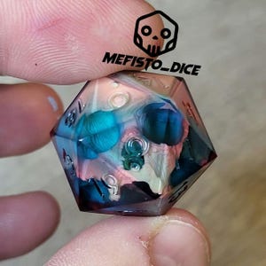 Può includere: Un dado di resina trasparente multicolore con un design a forma di teschio. Il dado presenta un motivo a spirale blu, verde, rosa e bianco. I numeri sul dado sono visibili. Il dado è etichettato "Mefisto_Dice".
