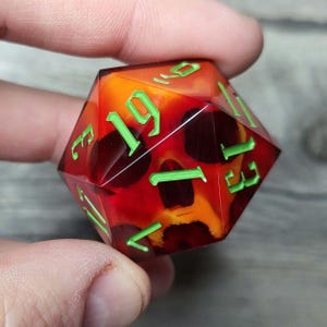 Può includere: Un dado da 20 facce rosso e arancione con numeri verdi. Il dado ha un design a forma di teschio su un lato.