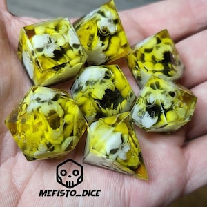 May include: Set of seven yellow and white dice with skull and bone designs. The dice are translucent and have a speckled appearance. The dice are on a hand.