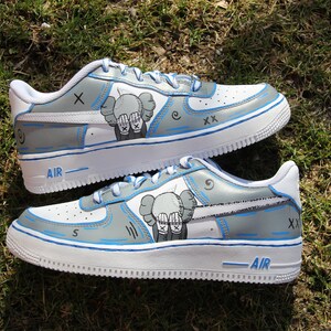 K&ouml;nnte beinhalten: Wei&szlig;e Low-Top-Sneaker mit blauen Akzenten und einer grauen und wei&szlig;en Illustration eines Charakters mit den H&auml;nden vor den Augen. Der Text "AIR" ist auf der Sohle des Schuhs sichtbar.