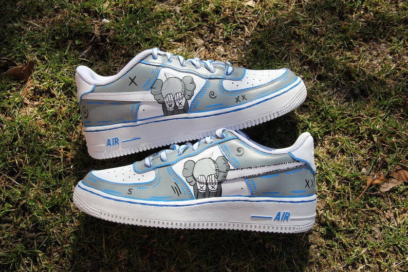 K&ouml;nnte beinhalten: Wei&szlig;e Low-Top-Sneaker mit blauen Akzenten und einer grauen und wei&szlig;en Illustration eines Charakters mit den H&auml;nden vor den Augen. Der Text "AIR" ist auf der Sohle des Schuhs sichtbar.