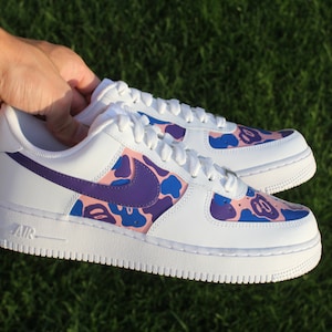 May include: White low-top sneakers with a purple swoosh and a pink and blue camouflage pattern.