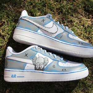 K&ouml;nnte beinhalten: Individuelle bemalte wei&szlig;e Nike Air Force 1 Sneaker mit blauen Akzenten. Die Sneaker haben ein graues und wei&szlig;es Design mit einer Comicfigur auf einem Schuh. Der Text "AIR" ist auf der Sohle sichtbar.