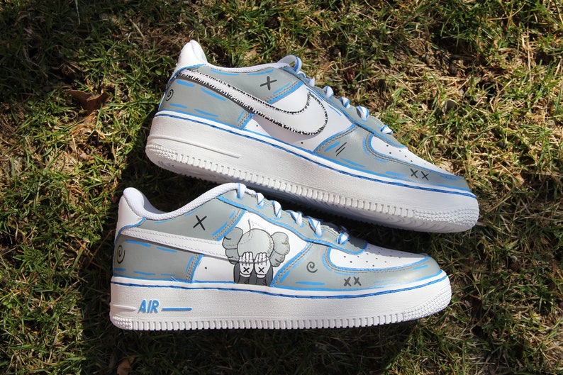 K&ouml;nnte beinhalten: Individuelle bemalte wei&szlig;e Nike Air Force 1 Sneaker mit blauen Akzenten. Die Sneaker haben ein graues und wei&szlig;es Design mit einer Comicfigur auf einem Schuh. Der Text "AIR" ist auf der Sohle sichtbar.