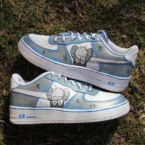 K&ouml;nnte beinhalten: Ein Paar wei&szlig;e Sneaker mit blauen Akzenten und grauem Hintergrund. Die Sneaker haben auf jeder Seite eine Comicfigur, mit dem Text "AIR" auf der R&uuml;ckseite jedes Schuhs.