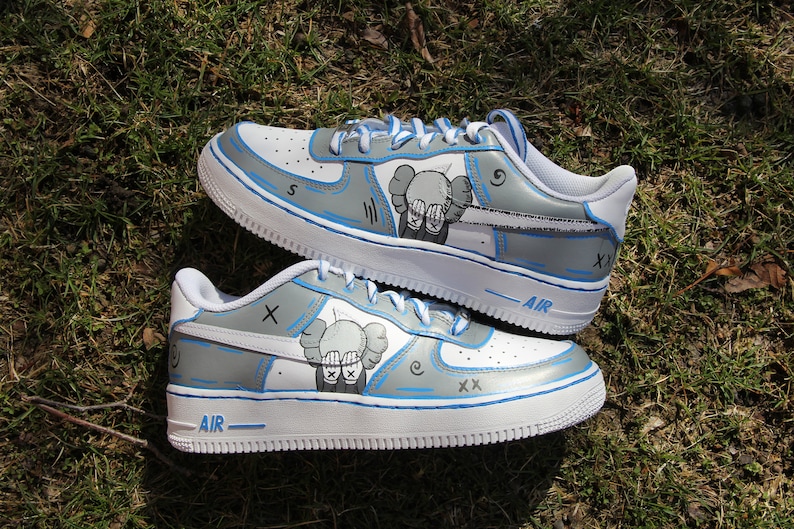 K&ouml;nnte beinhalten: Ein Paar wei&szlig;e Sneaker mit blauen Akzenten und grauem Hintergrund. Die Sneaker haben auf jeder Seite eine Comicfigur, mit dem Text "AIR" auf der R&uuml;ckseite jedes Schuhs.