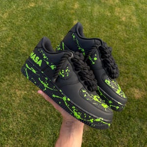 May include: Black sneakers with neon green paint splatter and the word "NASA" in neon green. The shoes have black rope laces and a black sole. The shoes are held up by a hand against a green grass background.