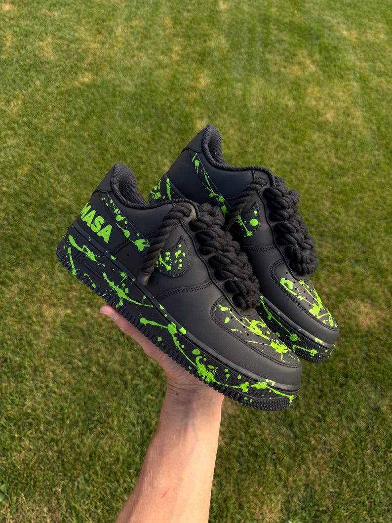May include: Black sneakers with neon green paint splatter and the word "NASA" in neon green. The shoes have black rope laces and a black sole. The shoes are held up by a hand against a green grass background.